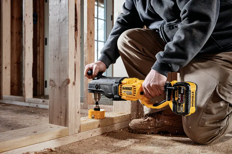 Uma pessoa está ajoelhada em um canteiro de obras usando uma furadeira sem fio DEWALT DCD470X1 com acessório de serra copo para perfurar um quadro de madeira.